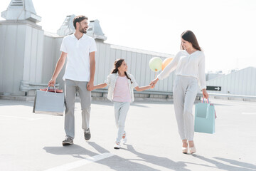 Family, father, mother and daughter are walking on road near parking after shopping in mall.