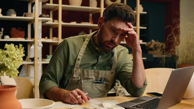 Upset confused worried Caucasian man male small business owner businessman potter counting expense bill debt bankrupt laptop shrugging shoulders gesture looking at camera worry indoors pottery store