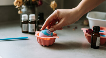 Hand molds a colorful bath bomb in a kitchen setting with essential oils and dried flowers in the background