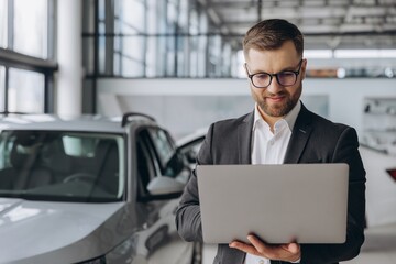 Modern bearded man in glasses and suit vehicle sales consultant using laptop inside car dealership