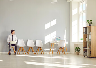 Young man sits on chair with laptop in an empty office, waiting a job interview. The atmosphere...