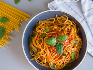 Balanesi pasta served in a blue bowl with fresh basil leaves and rich tomato sauce, accompanied by raw spaghetti and basil leaves on a gray table with a white striped napkin