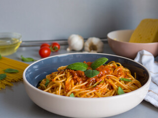 Balanesi pasta served with tomato sauce, basil leaves, and raw ingredients such as garlic, tomatoes, cheese, and olive oil are arranged on a gray table