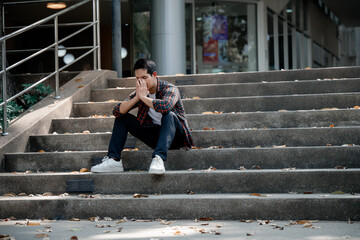 Exhausted college student sitting on campus stairs, hands covering face, experiencing overwhelming...