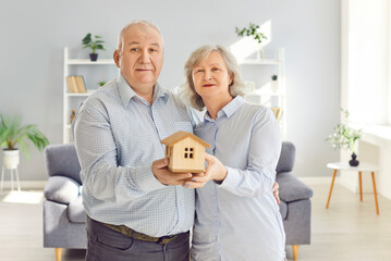 Happy senior couple holding wooden toy house in hands standing in living room at home looking at...