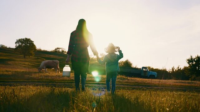 Morning in farm, adult woman and little son walking in field with grazing cow . Organic farming in ecological region, simple life and enjoy nature, back view of silhouette mother and child in sun