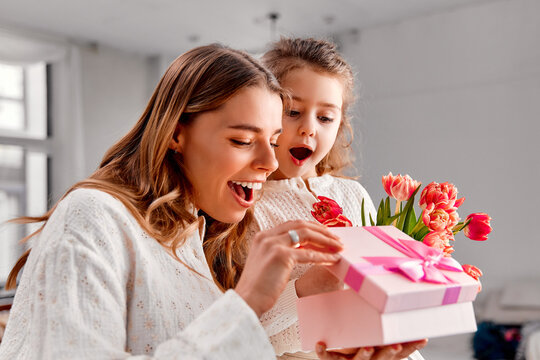 Happy Mother's Day! A happy mother and her excited daughter open a pink gift box together. The mother holds a bouquet of tulips while both smile with delight in a bright, cozy home setting.