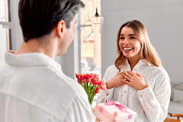 A man is congratulating his wife by giving her a bouquet of flowers and a gift. His excited and happy wife is rejoicing. The loving couple is at home in a bright apartment.