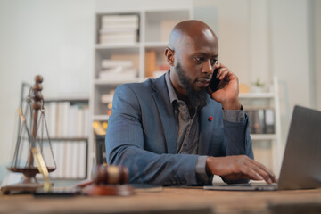Black lawyer wearing blue suit working on laptop and talking on cell phone while sitting at desk in...