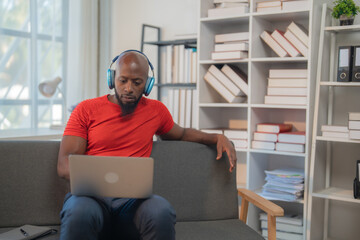 Young black man wearing headphones and using laptop while working remotely from home, sitting comfortably on a sofa in his living room or home office, surrounded by books and documents