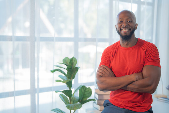 Portrait of a smiling african american man standing with arms crossed in a home office, exuding confidence and professionalism with a bright window and houseplant in the background