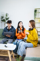 Three Asian young adult students sit in university living room, discussing their study abroad experiences, academic research, preparing thesis report, planning future global education opportunities.
