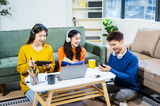 Three Asian young adult students sit in university living room, discussing their study abroad experiences, academic research, preparing thesis report, planning future global education opportunities.