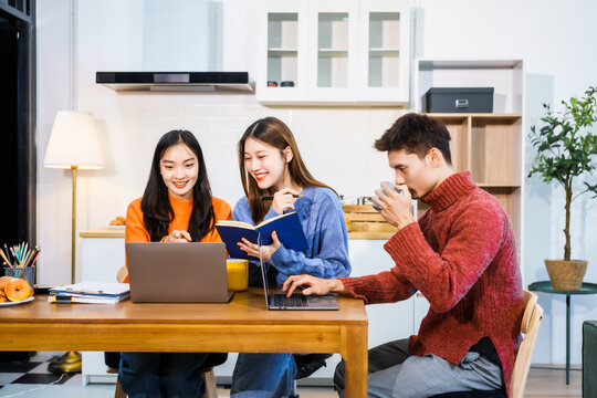 Three Asian young adult students sit around the kitchen table, attending online class on tablet. They engage in video conferencing, discussing lessons, taking notes, digital learning experience.
