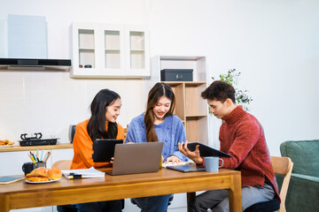 Three Asian young adult students sit around the kitchen table, attending online class on tablet....