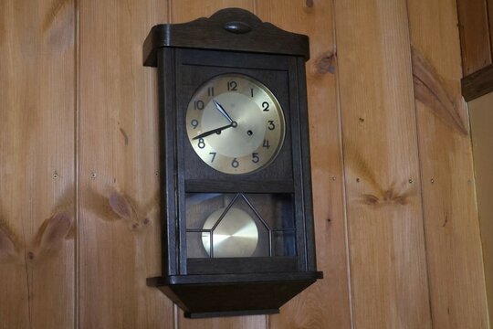 A classic vintage wooden wall clock with a brass dial and pendulum, mounted on a wooden-paneled wall. Timeless rustic home decor