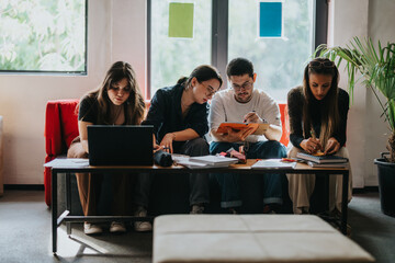 A group of four students works together on school tasks in a cozy study area. They are engaged with laptops, tablets, and notebooks, fostering teamwork and collaboration.