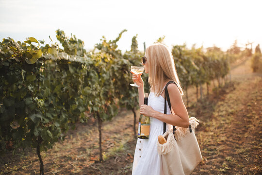 Stylish woman wearing white casual dress holding bottle of wine drinking from glass in vineyard at sunset. Autumn season.