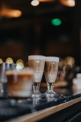A beautifully captured scene featuring coffee drinks in stylish glass mugs on a dark table, with warm bokeh effects in the background creating a cozy cafe atmosphere.