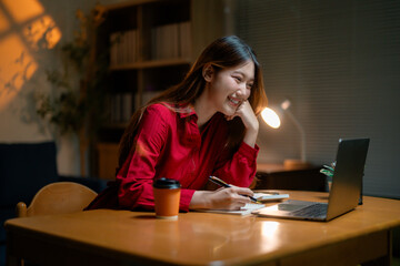 Professional Asian businesswoman wearing red shirt writing notes during late-night video conference, working remotely from home office with laptop