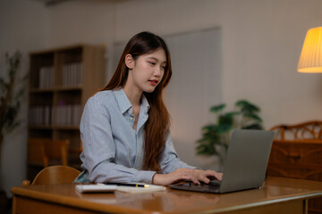 Focused female professional working late, typing on laptop at home office, surrounded by notes, reference books, soft lamp illumination highlighting workspace