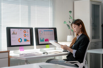 Professional Asian businesswoman reviewing financial documents, analyzing data on multiple screens with focused precision in contemporary corporate workspace