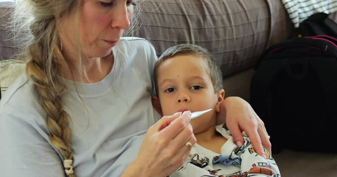 Blonde mother takes thermometer from under son arm looking at results on sofa. Careful woman with braid hugs preschooler boy checking temperature