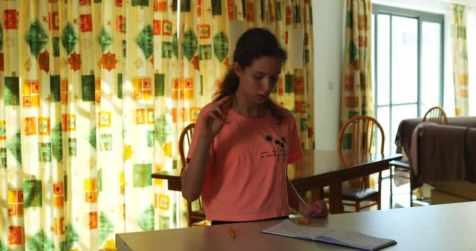 Studious woman focuses on notes performing sign language gestures. Diligent brunette recreates finger movements to be interpreter at table against window curtain in room