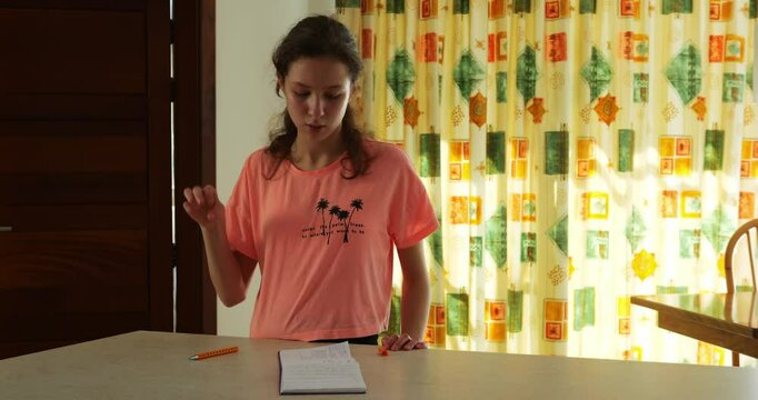 Mindful teenager intently looks at notepad while reproducing finger gestures. Long-haired young woman focuses on memos practicing hand motions to ne interpreter