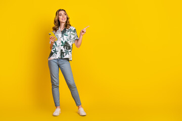 Happy young woman standing against yellow background holding smartphone and pointing, stylish in trendy printed shirt
