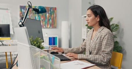 An attractive, experienced company accountant sits in office in front of a computer checking...