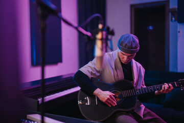 Obraz premium A musician focuses intently on playing the guitar while recording in a studio. The ambiance is modern, with a piano and microphones suggesting a creative music production environment.