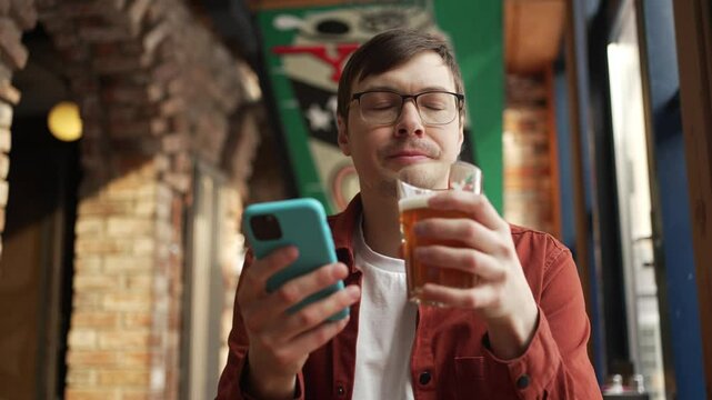 Young man wearing glasses enjoying beer while browsing smartphone, sitting in cozy pub with brick wall and soft ambient lighting, representing modern urban leisure lifestyle