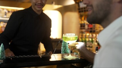 Special drink in Midori Sour cocktail with caramelized citrus slice and cinnamon power served stylish saucer glass by bartender serving to smart guest at luxurious bar christmas tree theme. Vinosity.