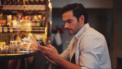 Smart man drinking cocktail in Old fashioned waiting friends or couple while chatting on smartphone to wait at counter bar with bartender preparing drinks at nightclub Friday meeting night. Vinosity.