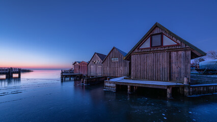 The old wooden boat sheds during the winter break in the blue hour before sunrise. The frozen ice...