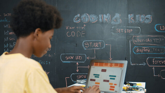 African boy using laptop programing system or coding prompt at blackboard with science theory. Highschool student working learning to write engineering code while sitting blackboard. Edification.
