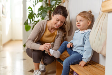 A young woman helps a little girl put on her shoes in a preschool or daycare dressing area, showing...
