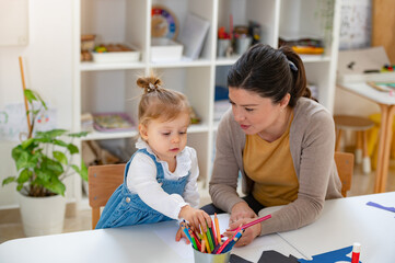 A young girl in a denim dress holds a colored pencil while interacting with her smiling teacher in...