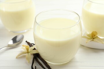 Delicious vanilla milk in glasses, pods and flowers on white wooden table, closeup
