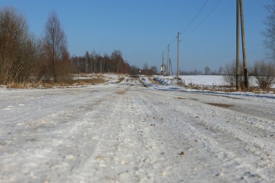 A winter countryside road covered in snow and ice, stretching into the distance. Power lines and bare trees frame the scenic rural landscape