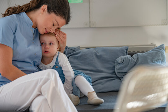 A caregiver in a blue uniform gently kisses a sick young girl's forehead while comforting her on a couch, showing warmth and care in a classroom or daycare setting