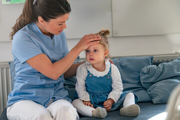 A concerned caregiver checks a young girl's forehead for fever as she sits on a couch in a classroom, looking unwell and in need of care and comfort