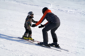Teaching a child to ski on alpine skis. A man backs up the baby on a snowy slope.