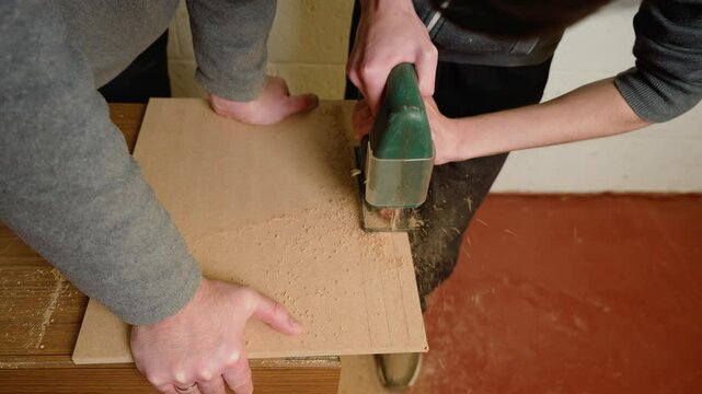 Father with son cutting wooden board with jigsaw in workshop. Dad holding board steady while boy operating power tool. Parent and child collaborating on carpentry project in hands-on learning activity