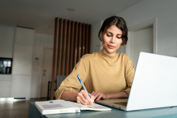Latin freelance businesswoman woman entrepreneur using computer for online remote business studying writing notes. Serious concentrated young hispanic female girl student working on laptop pc at home