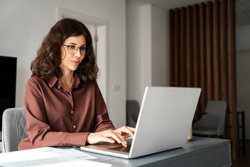 Focused young adult female freelancer financial specialist in eyeglasses typing on laptop computer,...