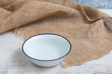 Vintage style empty white enamel bowl with black rim on white wooden table and jute fabric, isolated