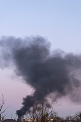 hick black smoke rising into the sky over residential buildings and trees at dusk. Environmental pollution and fire disaster concept. 