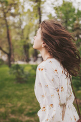 Fototapeta premium A young woman with flowing hair enjoys the warmth of a sunny day in a vibrant park. Dressed in a floral dress, she savors the tranquility of nature, letting the breeze lift her spirits